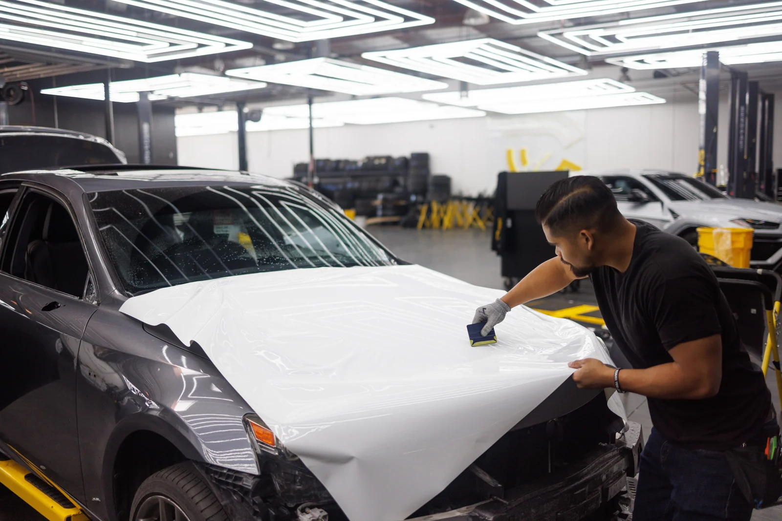 Vehicle wrap installer applying white vinyl to a car hood in a workshop